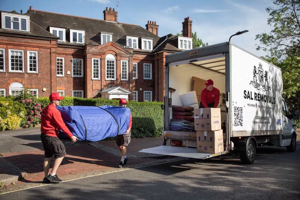 Two movers loading a blue blanket-wrapped sofa into a Luton van while a third stacks boxes