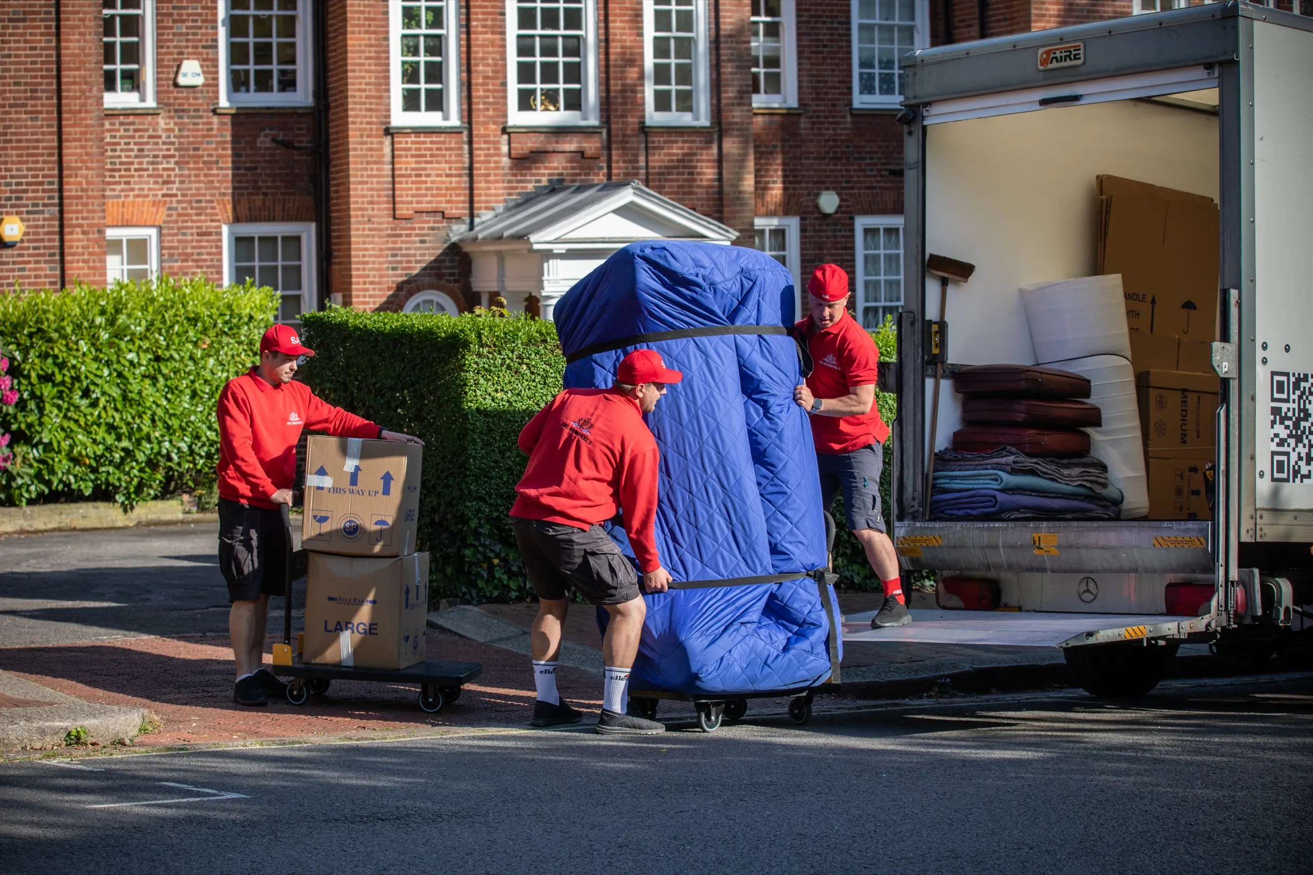 Two of the crew wheeling a wrapped sofa along a red-brick street